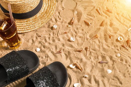 Summer beachwear, flip flops, hat, cold drink in a glass and seashells on sand beach. Top view. Copy space. Sun flare