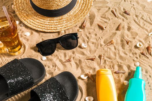 Summer beachwear, flip flops, hat, cold drink in a glass and seashells on sand beach. Top view. Copy space