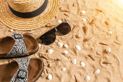 Summer beachwear, flip flops, hat, sunglasses and seashells on sand beach. Top view. Copy space. Sun flare