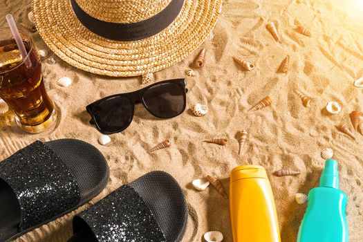 Summer beachwear, flip flops, hat, cold drink in a glass and seashells on sand beach. Top view. Copy space. Sun flare