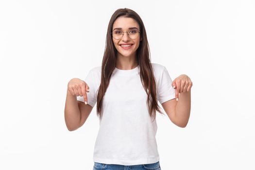 Click here. Portrait of brunette gorgeous young woman in glasses, pointing fingers down to show advertisement, smiling pleased, suggest visit link, recommend download or subscribe, white background.