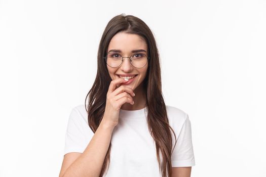 Close-up portrait of excited feminine, cute young woman in glasses, smiling and looking with temptation or desire, touching lip, want try something tasty, standing white background.