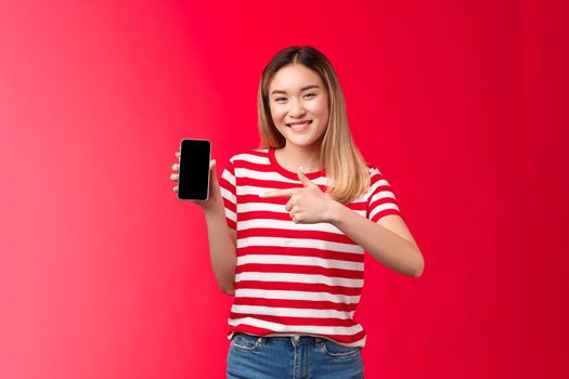 Very satisfied cheerful cute asian blond girl smiling broadly show smartphone display, pointing phone screen pleased introduce awesome new app, recommend use application, red background.