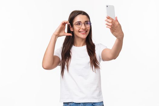 Communication, technology and lifestyle concept. Portrait of charming smiling young woman in glasses, taking selfie on mobile phone trying make pretty cute photo for social media.