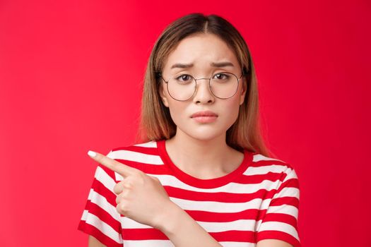 Sad cute asian female coworker tired working wear glasses frowning, pointing left look camera upset drained feel pressured distressed, stand red background unhappy wear striped t-shirt.