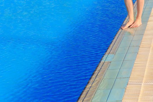 Young woman legs standing on stone border in front of swimming pool with blue clean water on a summer day.