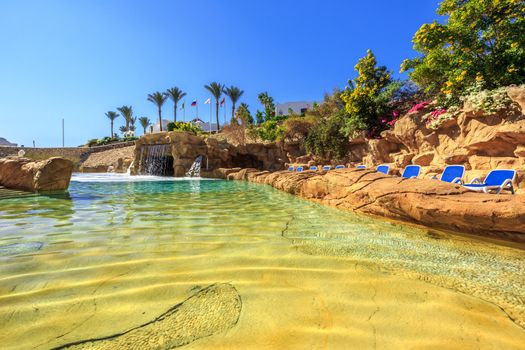 Swimming pool with artificial waterfall and sun loungers, luxury buildings, palm trees, behind it on a beautiful resort in Egypt