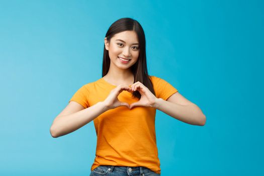 Lovely caring attractive asian girlfriend show heart sign cherish and value relationship, smiling broadly supportive, stand blue background wear yellow t-shirt, promote peace and happiness.