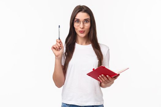 Creative enthusiastic young woman have great idea, writing it down in her notebook not to forget, raising pen in eureka sign, smiling excited, prepare plan or daily schedule, white background.