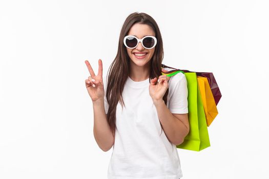 Shopping, leisure and lifestyle concept. Relaxed, carefree attractive modern feminine girl, holding shop bags behind shoulder and make peace sign, wear sunglasses, buying clothes for summer.