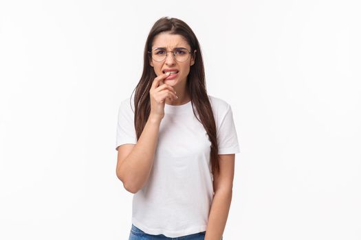 Waist-up portrait of embarrassed young cute girl feel nervous, squinting and biting lip anxious, afraid someone realise what she did, standing concerned, alarmed over white background.