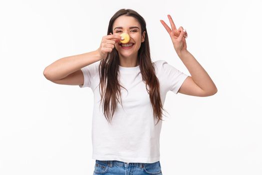 Portrait of positive, cheerful young girl eating sweets, holding macaron over nose, make peace sign, look kawaii and silly, laughing joyful, visit favorite dessert cafe, white background.