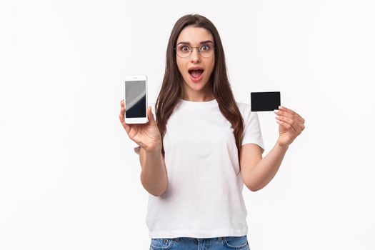 Portrait of excited and happy young woman introduce new application, favorite online store to buy clothes, show mobile phone and credit card, smiling amused, white background.