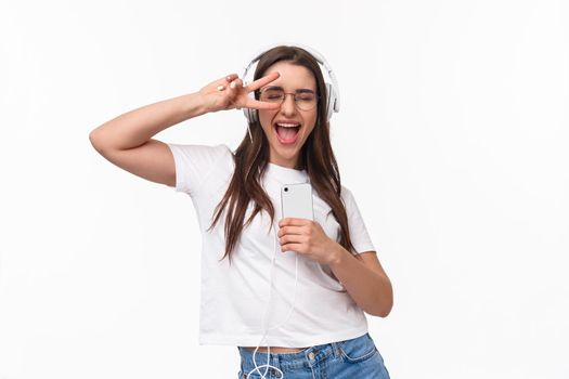 Technology, lifestyle and music concept. Carefree excited happy woman dancing, vibing and enjoying favorite songs in headphones, close eyes show peace sign, holding smartphone, white background.
