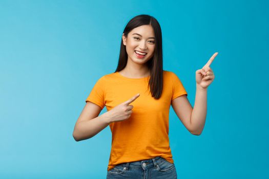 Cheerful lively cute asian woman smiling laughing upbeat, pointing upper right corner, give advice what choose, shopping online, grinning happily, enthusiastic promo, blue background.