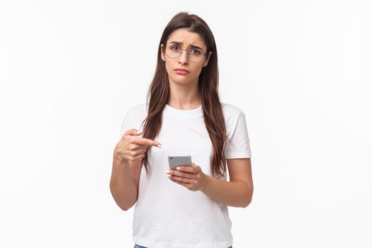 Communication, technology and lifestyle concept. Waist-up potrait of sad and gloomy young uneasy caucasian woman in glasses, frowning and pointing at mobile phone with regret or sadness.