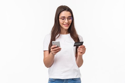 Portrait of attractive young woman in glasses holding credit card and smartphone, enter digit numbers of her banking account to shopping application, order online, stand white background.