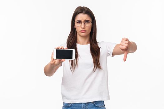 Communication, technology and lifestyle concept. Portrait of upset and disappointed young female rated bad app, showing content on mobile phone, grimacing dislike, show thumbs-down disapproval.