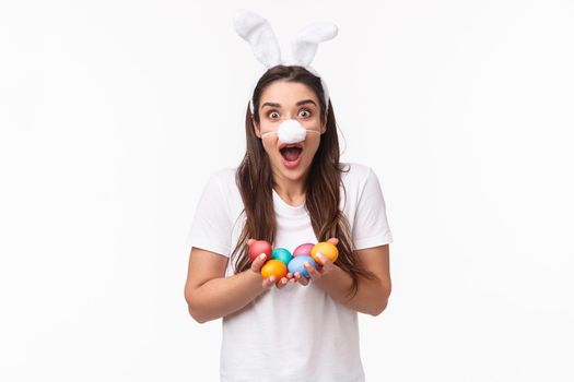 Portrait of excited happy young funny woman wearing rabbit costume, ears and fluffy nose mask, holding colored eggs, celebrating holiday Easter party, stand amazed white background.