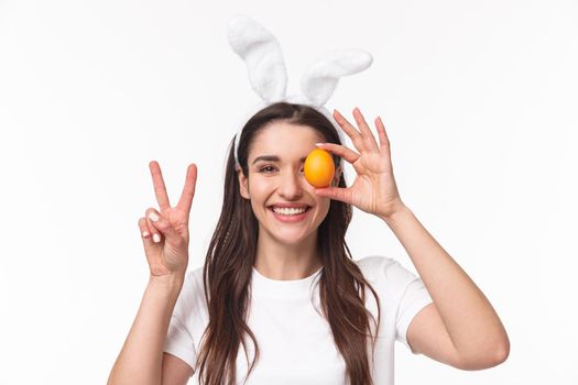 Easter, holidays and spring concept. Close-up portrait of cheerful, upbeat young woman in rabbit ears, bragging with her first colored egg on Easter day, smiling and showing peace sign.