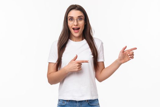 Waist-up portrait of excited, enthusiastic brunette 25s woman in t-shirt, jeans, pointing fingers right and look astonished with amused smile at camera, suggesting visit awesome new place.