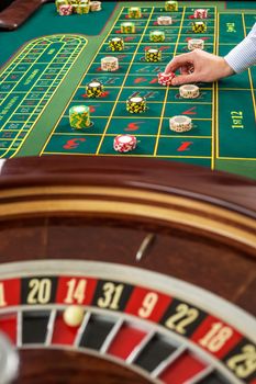 Roulette and piles of gambling chips on a green table in casino. Man hand over casino chips on roulette table