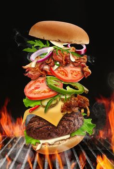 Big hamburger with flying ingredients roasted on barbecue BBQ grill with bright flaming fire against black background. Beef cutlet, ham, cheese, vegetables and greens. Cooking, fast food. Close up
