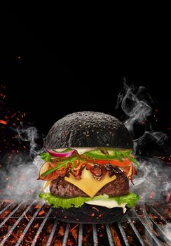 Black burger roasted on barbecue BBQ grill with flaming fire, sparks and smoke against black background. Beef cutlet, ham, cheese, vegetables and greens. Cooking, fast food. Close up, copy space