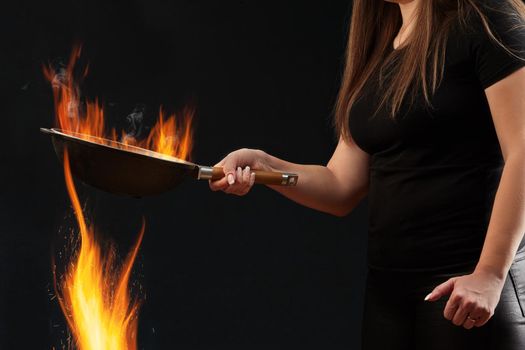 Brunette girl with tattooed hands and long hair, dressed in leggings and t-shirt. Holding burning wok pan above fire against black studio background. Cooking concept. Close up, copy space, side view