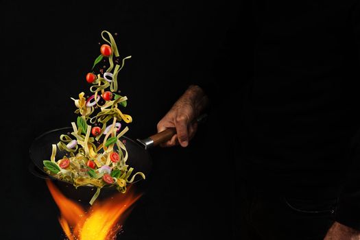 Unrecognizable man dressed in jeans is holding wok pan above fire and cooking pasta with cherry tomatoes, onion and basil against black studio background. Close up, copy space