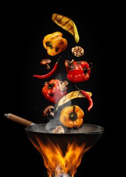 Wok pan with smoke above fire is frying yellow and red bell pepper, hot pepper, mushrooms, zucchini, garlic against black studio background. Cooking concept. Close up, copy space