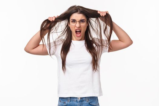 Waist-up portrait of bothered and annoyed, angry pissed-off young girlfriend losing temper, pulling hair from head and screaming being furious and outraged, white background.