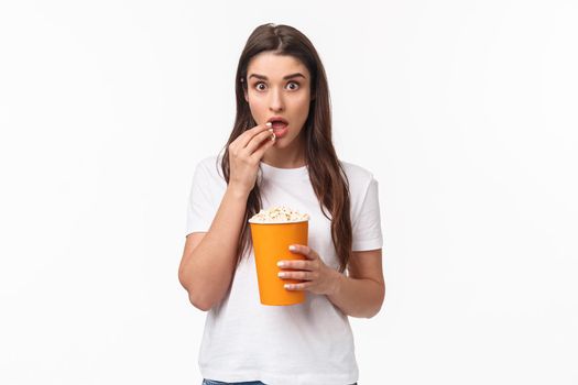 Entertainment, fun and holidays concept. Portrait of woman entertained, watching movie with interest, enjoying favorite tv series, eating popcorn and staring camera, stand white background.