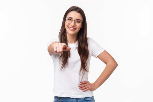 Waist-up portrait of happy smiling young woman inviting you join her team, recruit person, pointing finger at camera grinning and standing confident in her choice, white background.
