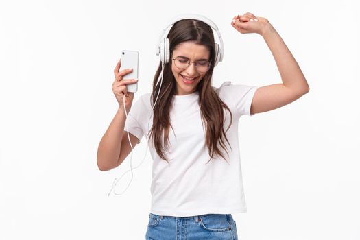 Technology, lifestyle and music concept. Portrait of excited and carefree relaxed beautiful woman having fun listening songs and playlist, vibing dancing alone in headphones, hold mobile phone.
