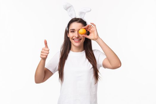Portrait of satisfied, happy young woman in rabbit ears, holding one colored eggs over eye, show thumbs-up painting it for Easter holiday party, celebrating traditional event, white background.