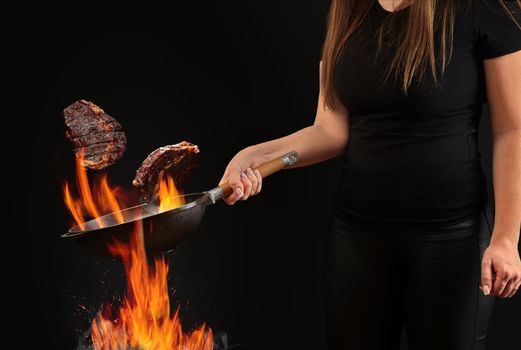 Brunette cook with tattooed hands, dressed in leggings and t-shirt. Holding wok pan above fire and frying two beef steaks against black background. Cooking concept. Close up, copy space, side view
