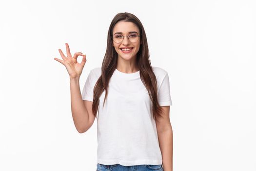 Waist-up portrait of optimistic, smiling happy young pretty woman in glasses, showing okay sign, agree or recommend something, give positive review, approve excellent choice, white background.