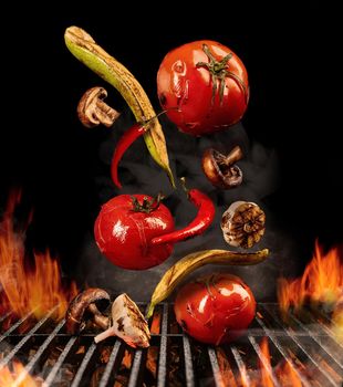 Grilled tomatoes, zucchini, chili, champignons, halves of garlic are falling down on black background. Barbecue bbq grill, flaming fire, ember charcoal, smoke. Cooking concept. Close up, copy space