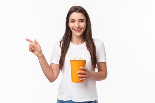 Entertainment, fun and holidays concept. Portrait of cheerful cute young girl waiting for friend in cinema, movie starting soon, pointing finger left inviting come join her, smiling, hold popcorn.