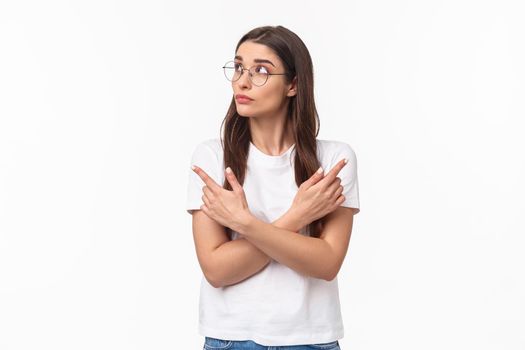 Waist-up portrait of indecisive thoughtful young woman in glasses, 20s female student picking her path, pointing fingers sideways left and right, making decision, thinking over white background.