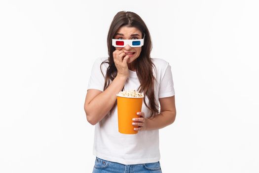 Entertainment, fun and holidays concept. Portrait of scared young cute girl in 3d glasses, eating popcorn, stooping and biting fingers as being scared of horror movie watching in cinema.