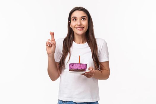 Entertainment, fun and holidays concept. Portrait of positive, smiling wishful young girl, cross finger good luck, bite lip eager wish come true, smiling up at blowing candle on birthday cake.