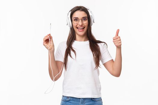 Waist-up portrait of excited, smiling happy and enthusiastic young girl in headphones, holding wire of earphones and show thumbs-up, ready to plug in to smartphone and listen some music.