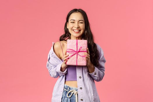 Portrait of cheerful, happy asian girl receive gift, smiling excited, holding wrapped present and cant hide happiness, celebrating birthday, being invited to b-day party, pink background.