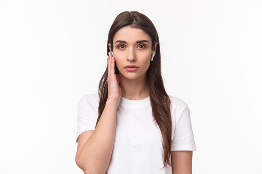 Close-up portrait of serious-looking young confident female asnwering calls using wireless headphones, touching earbud to change volume or answer call, look camera determined, white background.