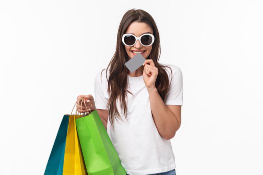 Shopping, leisure and lifestyle concept. Portrait of excited beautiful young woman cant wait to waste all her money, biting credit card and look thrilled, think what store visit next, hold shop bags.