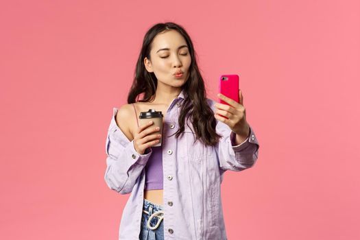 Portrait of stylish good-looking female blogger, lifestyle internet influencer taking selfie with take-away coffee from her favorite cafe, hold mobile phone, pouting for kiss and close eyes.
