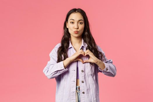Portrait of romantic cute korean girl show heart gesture and fold lips, giving kiss, express sympathy, passion and romance, standing pink background in lovely stylish outfit.