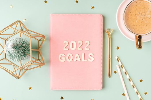 Top view Desktop Christmas pink notepad with 2022 goals letters text. Flat lay on green mint table background with planner, cup of coffee, candle, Christmas decoration, notebook and stationery.